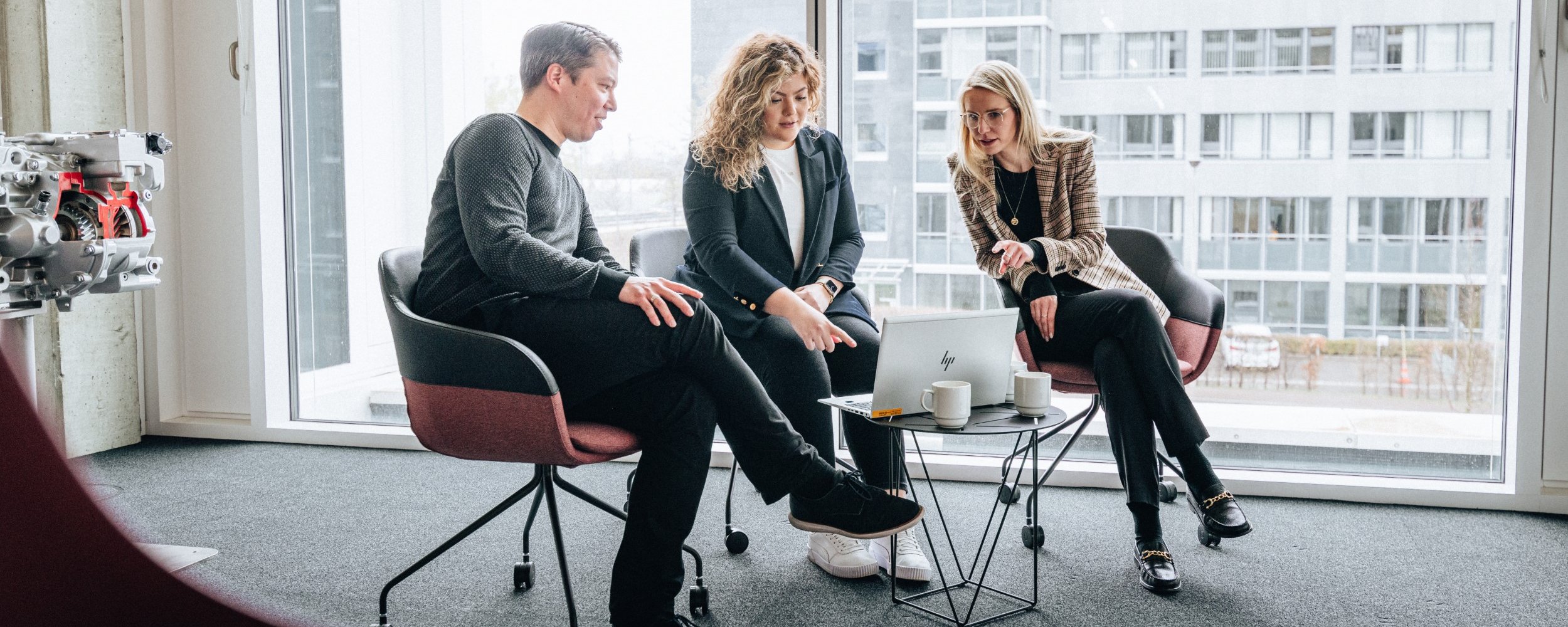 Three people are sitting in a brightly lit room on the Daimler Truck Campus, discussing something on a laptop placed on a small table.