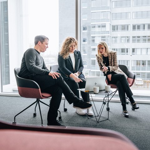 Three people are sitting in a brightly lit room on the Daimler Truck Campus, discussing something on a laptop placed on a small table.