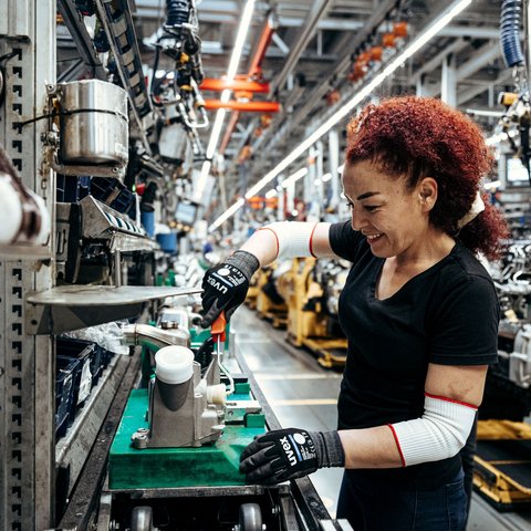 A red-haired woman wearing safety gloves works on a Daimler Truck production part in a production hall.