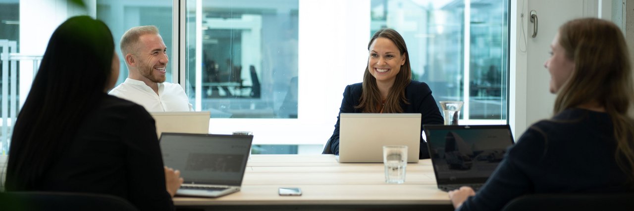 A meeting in a modern office where four employees are chatting in a relaxed atmosphere and taking notes on their laptops.