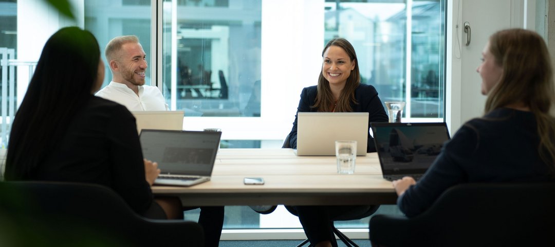 A meeting in a modern office where four employees are chatting in a relaxed atmosphere and taking notes on their laptops.