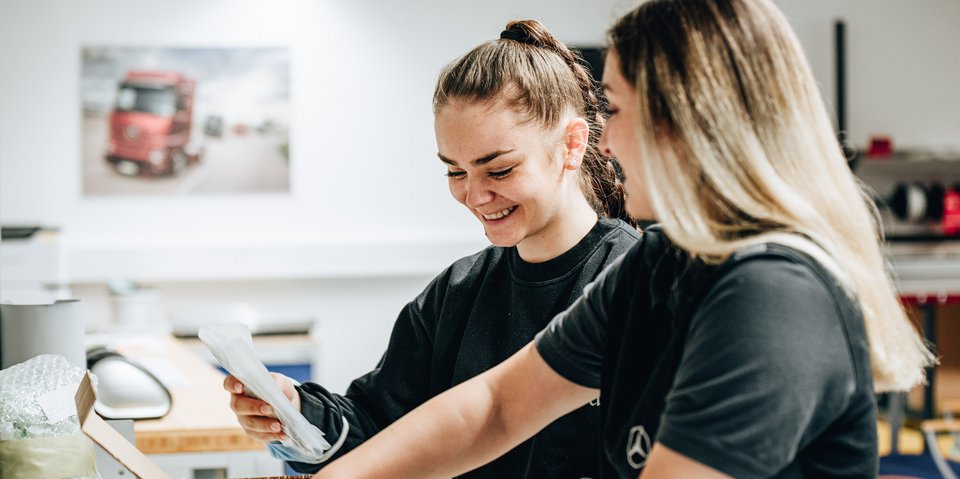 Two cheerful trainees in black work clothes pack a box together.