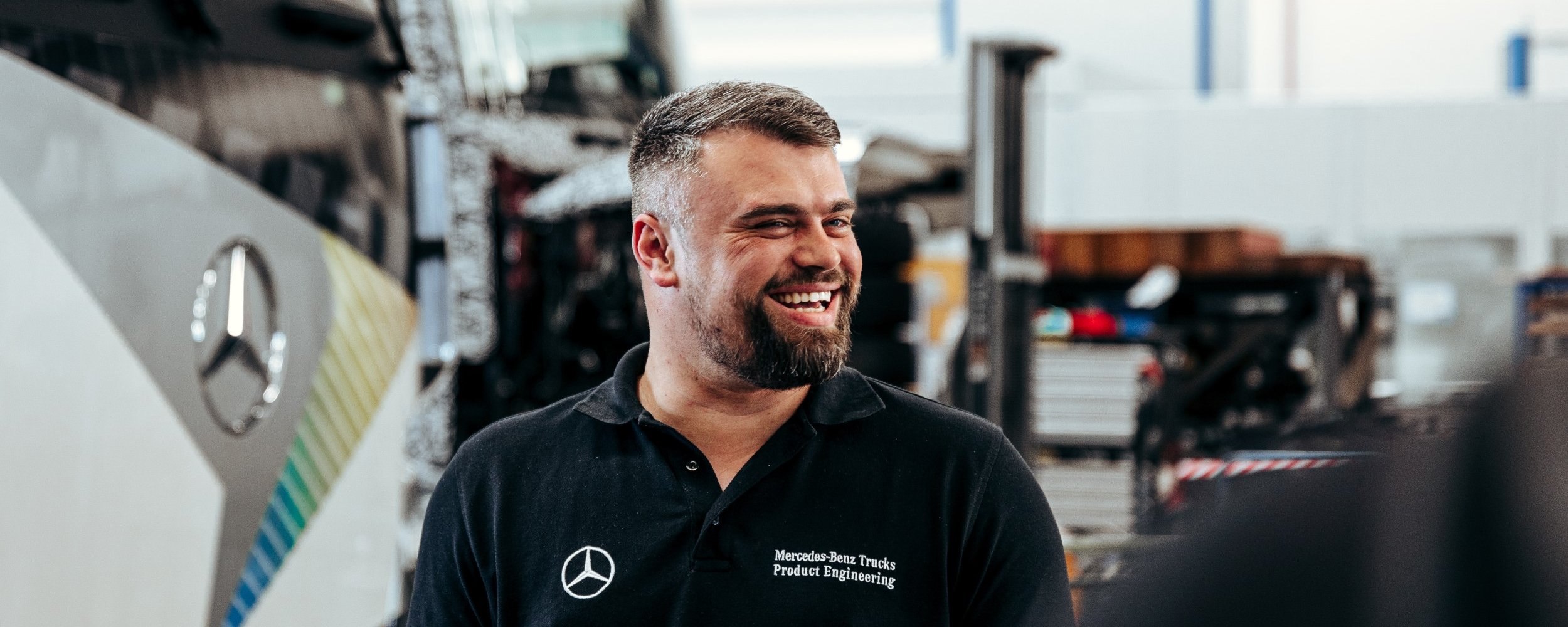 Employee in a black polo shirt smiles in a Mercedes-Benz Trucks workshop. A Daimler Truck lorry can be seen in the background.