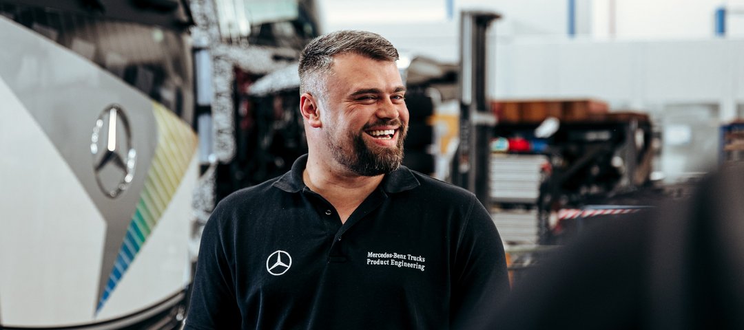 Employee in a black polo shirt smiles in a Mercedes-Benz Trucks workshop. A Daimler Truck lorry can be seen in the background.