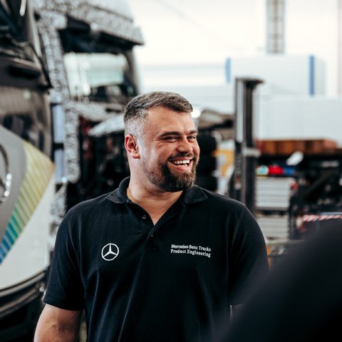 Employee in a black polo shirt smiles in a Mercedes-Benz Trucks workshop. A Daimler Truck lorry can be seen in the background.