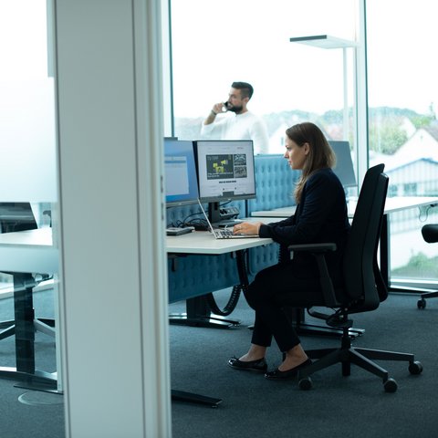 A woman in business attire works intently at two monitors in the Legal & Compliance department of Daimler Truck. A man is on the phone in the background.