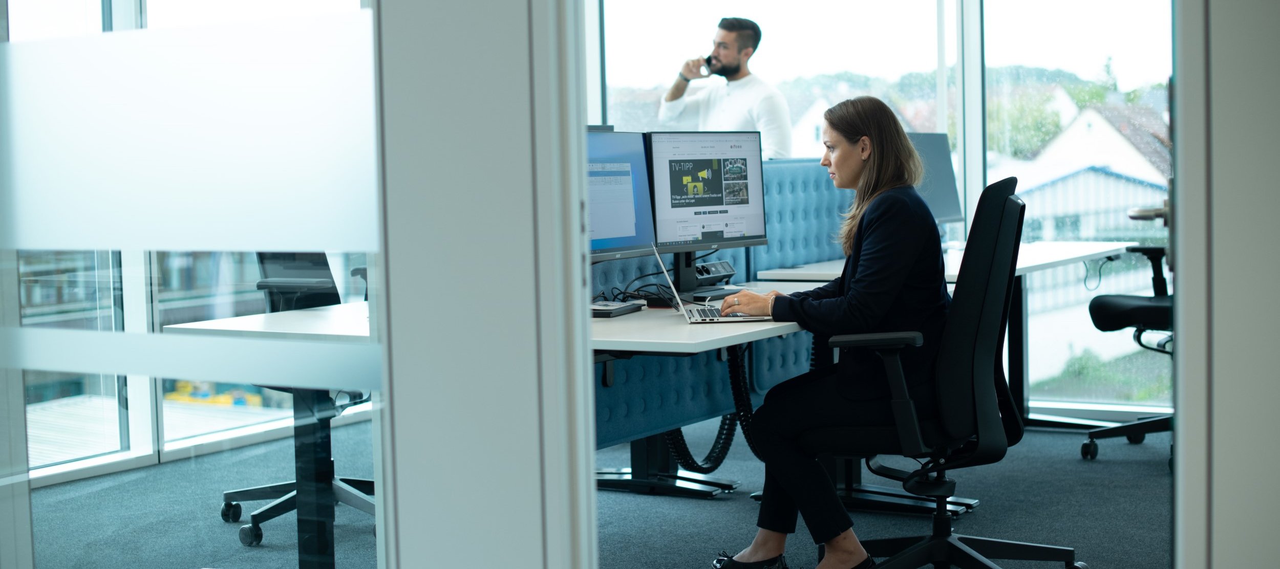 A woman in business attire works intently at two monitors in the Legal & Compliance department of Daimler Truck. A man is on the phone in the background.