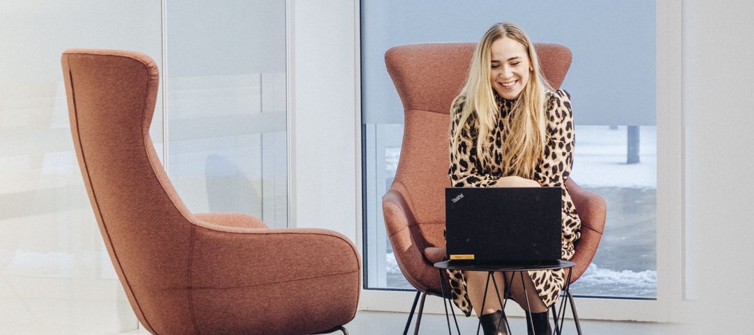 A young employee sits on an armchair in a modern, bright lounge area and works on a laptop on a small table in front of her.