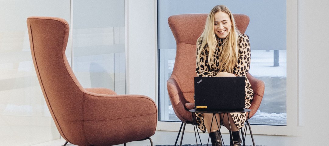 A young employee sits on an armchair in a modern, bright lounge area and works on a laptop on a small table in front of her.