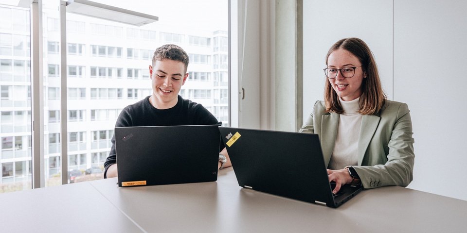 Two doctoral students, a young man and a young woman, sit next to each other and work on their laptops at the Daimler Truck Campus.