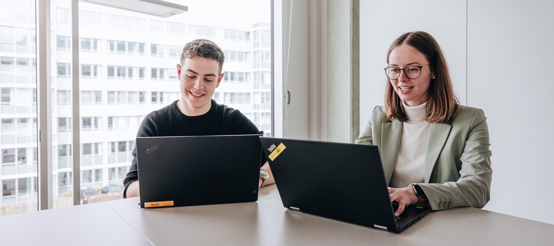 Two doctoral students, a young man and a young woman, sit next to each other and work on their laptops at the Daimler Truck Campus.