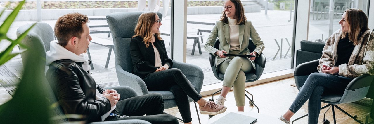 Four students sit on comfortable armchairs in a modern, light-flooded room and chat in a relaxed atmosphere, with an outdoor terrace in the background.