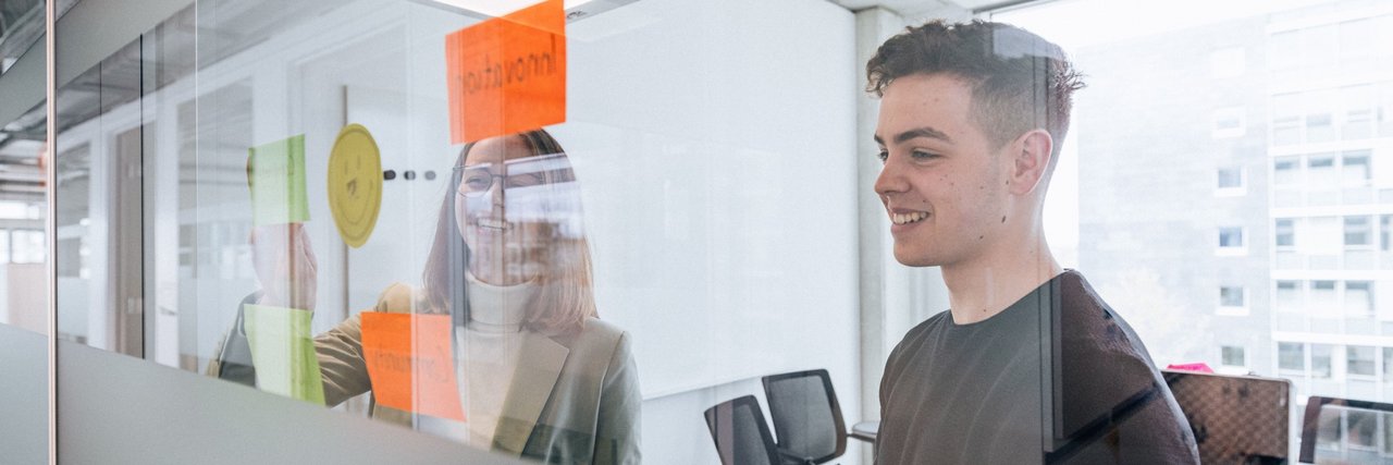 Two young employees stand behind a glass wall with colourful sticky notes, the woman smiles while the man arranges a note.