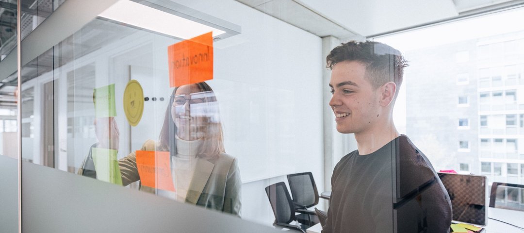 Two young employees stand behind a glass wall with colourful sticky notes, the woman smiles while the man arranges a note.