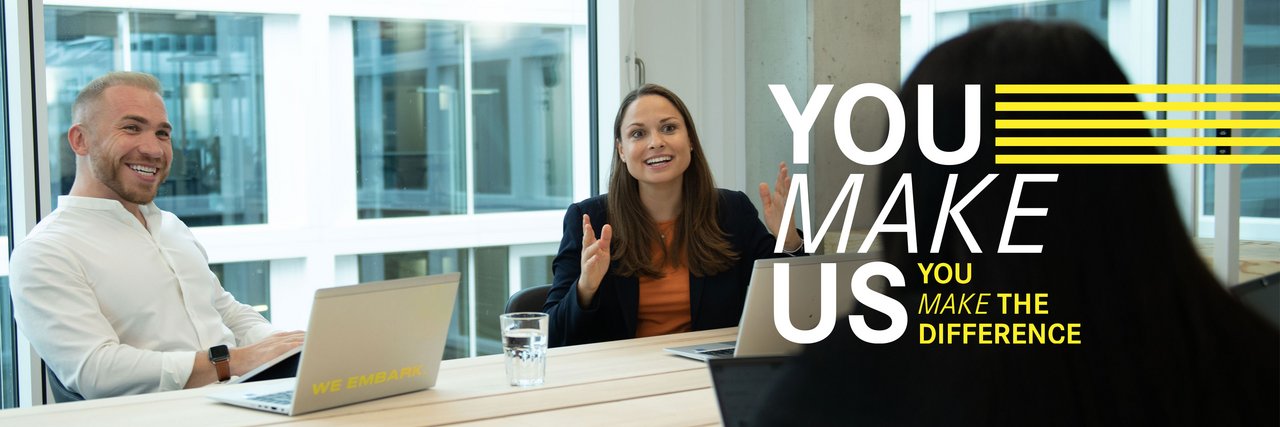 Three colleagues sit across from each other laughing at a meeting table with laptops. Above the picture is the "You Make Us" lettering.