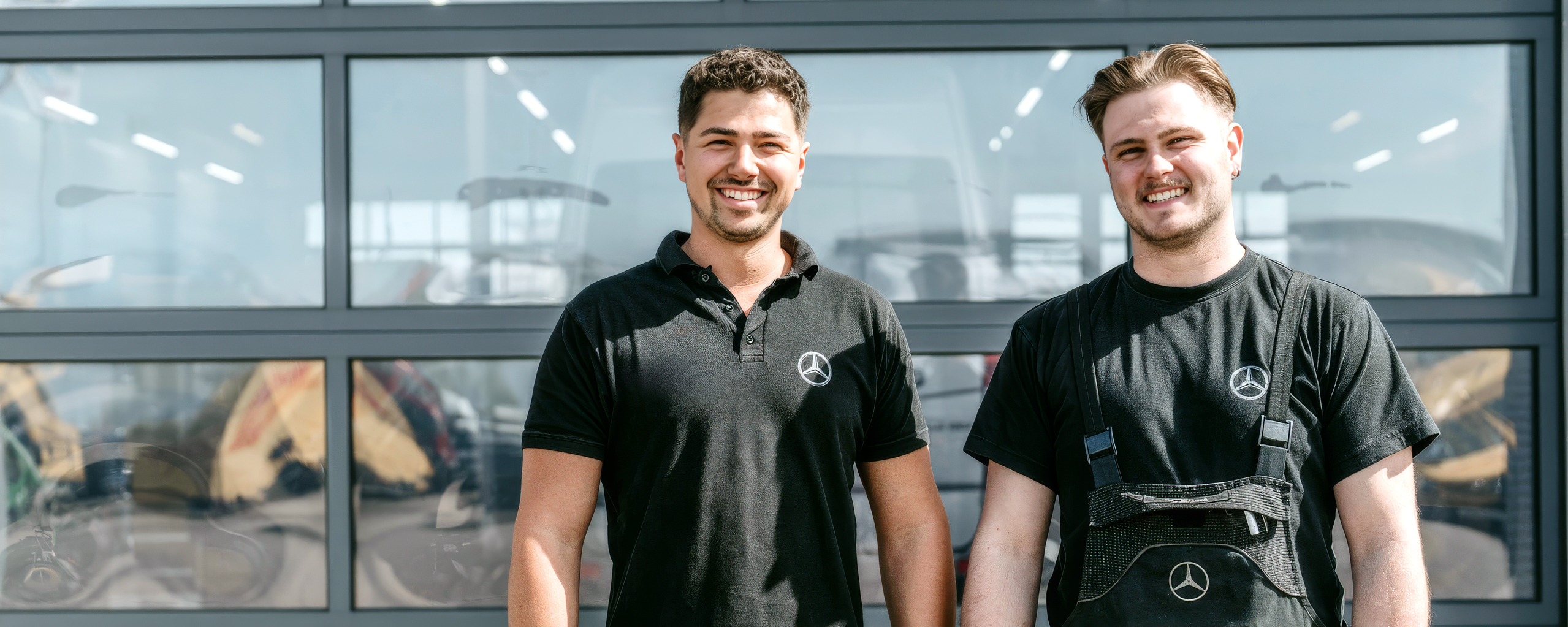 Two technicians from Daimler Truck's commercial vehicle centers stand smiling in front of a modern workshop building. Both are wearing black work clothes with the Mercedes-Benz logo.